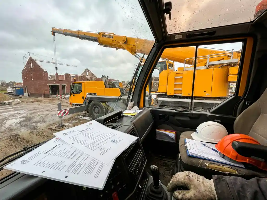Gele mobiele telescoopkraan op Nederlandse bouwplaats met vergunningsdocumenten op dashboard en veiligheidshelmen