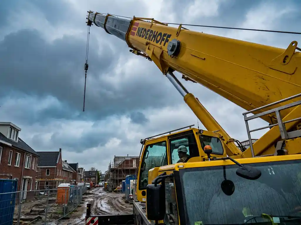 Gele mobiele kraan met uitgestrekte giek boven Nederlands bouwterrein, gecertificeerde machinist zichtbaar in cabine, bewolkte lucht.