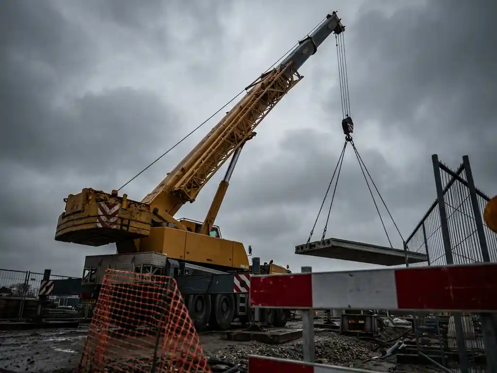 Mobiele kraan heft betonnen last aan strakgespannen stalen kabels op Nederlandse bouwplaats, veiligheidsbarrières op voorgrond, bewolkte lucht.
