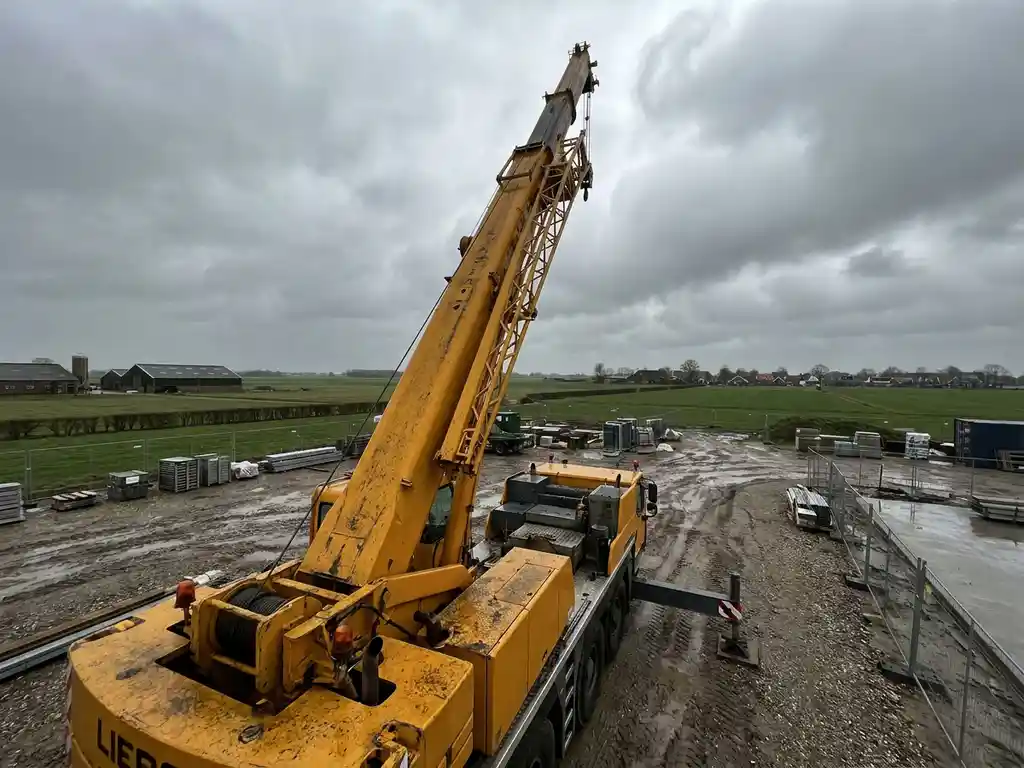 Gele vrachtwagenkraan met uitgestrekte giek op een bouwplaats in Limburg, omringd door vlak weiland onder bewolkte lucht.